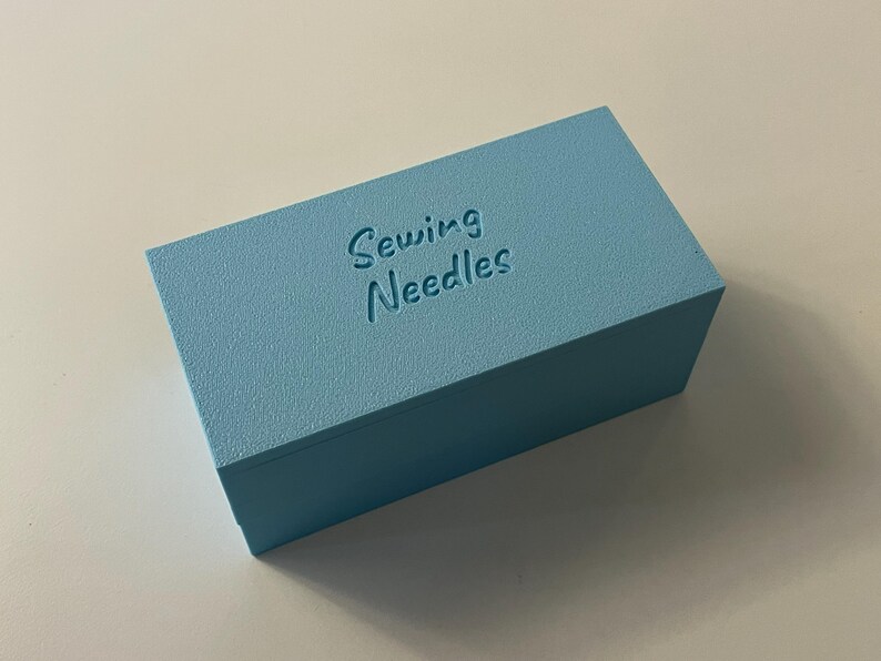 May include: A light blue rectangular box with the words "Sewing Needles" embossed on the top. The box has a textured surface and is sitting on a white surface. The box is a solid color.