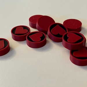 Puede incluir: Un conjunto de piezas de juego rojas con un dise&ntilde;o de coraz&oacute;n. Las piezas son circulares con un borde elevado y un interior negro. Algunas tienen forma de coraz&oacute;n en el centro, mientras que otras son discos rojos s&oacute;lidos.