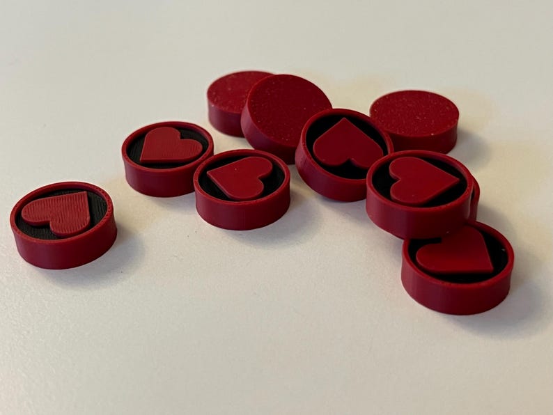 Puede incluir: Un conjunto de piezas de juego rojas con un dise&ntilde;o de coraz&oacute;n. Las piezas son circulares con un borde elevado y un interior negro. Algunas tienen forma de coraz&oacute;n en el centro, mientras que otras son discos rojos s&oacute;lidos.