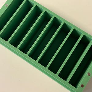 May include: A rectangular, green soap mold with ten equally spaced, rectangular compartments. Small, silver magnets are visible on the corners of the mold. The mold is designed for making bars of soap.