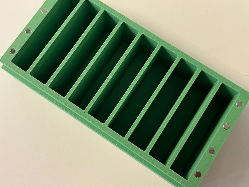 May include: A rectangular, green soap mold with ten equally spaced, rectangular compartments. Small, silver magnets are visible on the corners of the mold. The mold is designed for making bars of soap.