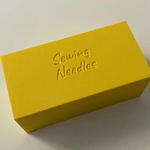 May include: A bright yellow rectangular box with the words "Sewing Needles" embossed on the top. The box is a solid color and appears to be made of a textured material. The box is likely used for storing sewing needles.