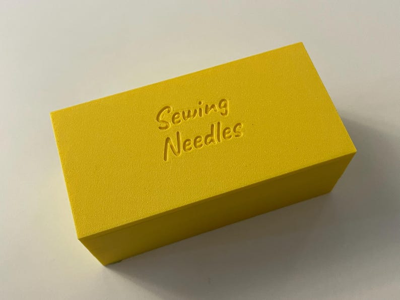 May include: A bright yellow rectangular box with the words "Sewing Needles" embossed on the top. The box is a solid color and appears to be made of a textured material. The box is likely used for storing sewing needles.