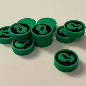 May include: A collection of green game tokens with a black center and a green lightbulb icon. The tokens are round and stacked, with some scattered on a white surface. The lightbulb icon is a prominent feature.