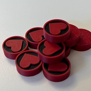Puede incluir: Un conjunto de fichas de juego redondas rojas con un dise&ntilde;o de coraz&oacute;n. Cada ficha tiene un centro negro y un coraz&oacute;n en relieve. Algunas fichas tienen una superficie texturizada. Est&aacute;n apiladas sobre una superficie blanca.