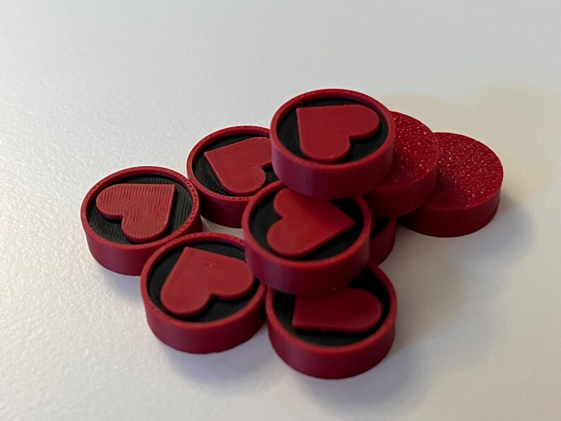 Puede incluir: Un conjunto de fichas de juego redondas rojas con un dise&ntilde;o de coraz&oacute;n. Cada ficha tiene un centro negro y un coraz&oacute;n en relieve. Algunas fichas tienen una superficie texturizada. Est&aacute;n apiladas sobre una superficie blanca.
