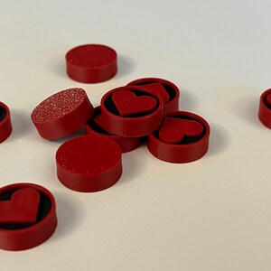 Puede incluir: Un conjunto de imanes rojos circulares con un dise&ntilde;o de coraz&oacute;n. Algunos imanes tienen una superficie roja s&oacute;lida, mientras que otros tienen una textura brillante. Los imanes est&aacute;n dispersos sobre una superficie blanca.