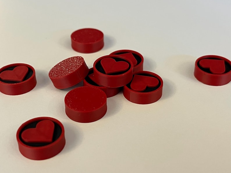 Puede incluir: Un conjunto de imanes rojos circulares con un dise&ntilde;o de coraz&oacute;n. Algunos imanes tienen una superficie roja s&oacute;lida, mientras que otros tienen una textura brillante. Los imanes est&aacute;n dispersos sobre una superficie blanca.