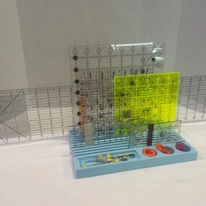 May include: A collection of quilting tools, including clear acrylic rulers with grid markings, a neon yellow square ruler, and a light blue organizer tray with scissors and other small tools. The rulers are marked in inches.