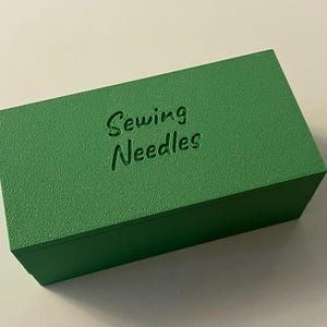 May include: A rectangular, textured, green box with the words "Sewing Needles" embossed on the top. The box is a solid, vibrant green color and appears to be made of a solid material.