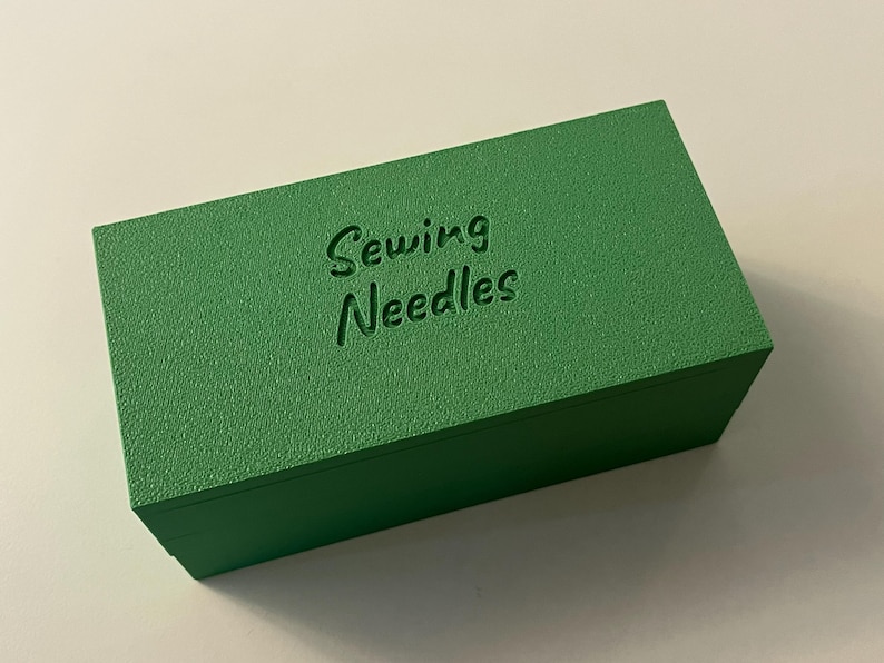 May include: A rectangular, textured, green box with the words "Sewing Needles" embossed on the top. The box is a solid, vibrant green color and appears to be made of a solid material.