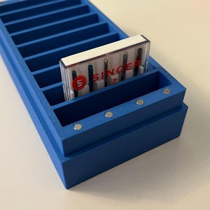 May include: A blue wooden sewing needle organizer with multiple compartments. A package of Singer sewing machine needles is placed in one of the compartments. The package has the Singer logo and the words "SINGER" printed on it.