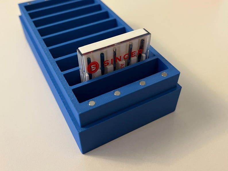 May include: A blue wooden sewing needle organizer with multiple compartments. A package of Singer sewing machine needles is placed in one of the compartments. The package has the Singer logo and the words "SINGER" printed on it.