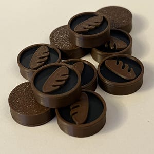 Puede incluir: Una colecci&oacute;n de fichas circulares marrones con un centro negro y un dise&ntilde;o de pan. Algunas fichas tienen una superficie texturizada. Las fichas est&aacute;n dispersas sobre una superficie de color claro.