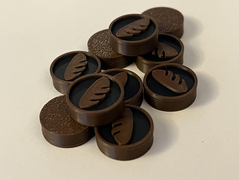 Puede incluir: Una colecci&oacute;n de fichas circulares marrones con un centro negro y un dise&ntilde;o de pan. Algunas fichas tienen una superficie texturizada. Las fichas est&aacute;n dispersas sobre una superficie de color claro.