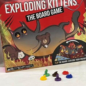 May include: The board game "Exploding Kittens" is displayed, featuring a red box with a cartoon cat illustration. The game includes colorful game pieces in various shapes and colors. The box text reads "The Board Game" and "Flip the board. Explode your friends."