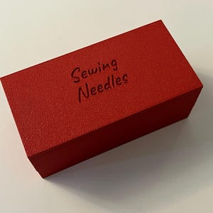 May include: A rectangular, red box with the words "Sewing Needles" embossed on the top. The box has a textured surface and is likely made of wood or a similar material. The box is sitting on a white surface.