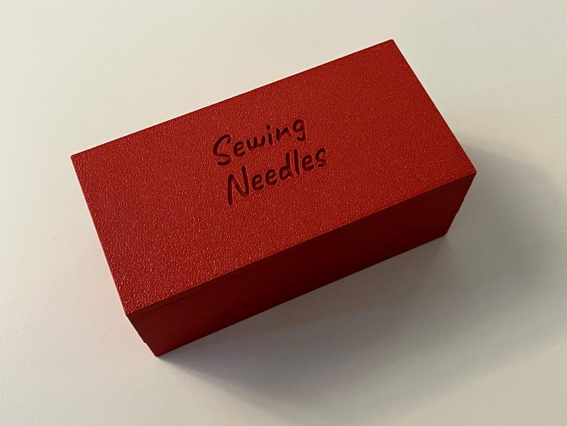 May include: A rectangular, red box with the words "Sewing Needles" embossed on the top. The box has a textured surface and is likely made of wood or a similar material. The box is sitting on a white surface.