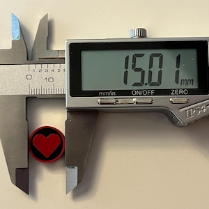 Puede incluir: Un calibrador digital mide un objeto peque&ntilde;o, redondo, rojo y negro con un dise&ntilde;o de coraz&oacute;n. La pantalla digital muestra 15,01 mm. El calibrador es plateado y gris.