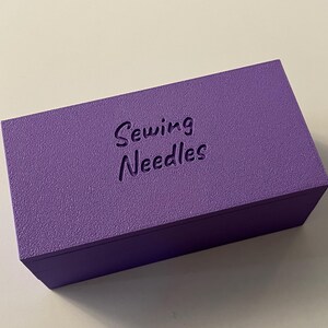 May include: A rectangular, textured, purple box with the words "Sewing Needles" embossed on the top. The box is a solid color and appears to be made of wood or a similar material. The box is designed for storing sewing needles.