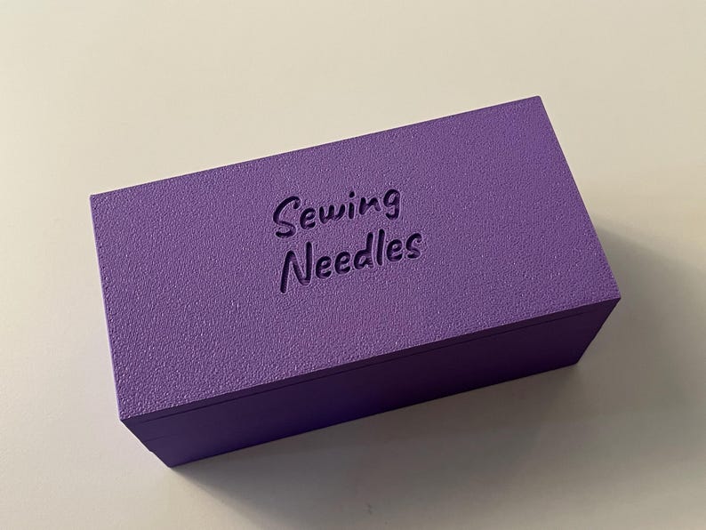 May include: A rectangular, textured, purple box with the words "Sewing Needles" embossed on the top. The box is a solid color and appears to be made of wood or a similar material. The box is designed for storing sewing needles.