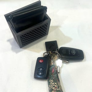 May include: A dark gray, geometric key and wallet organizer with a black leather wallet inside. A set of keys with a floral patterned keychain and two car key fobs are next to the organizer.