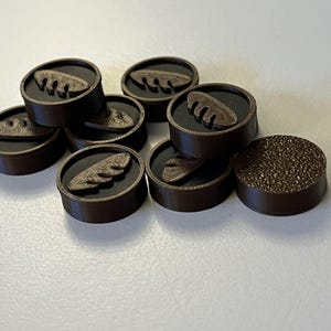 Puede incluir: Una colecci&oacute;n de fichas de juego circulares marrones. Cada pieza tiene un borde marr&oacute;n oscuro, un interior negro y un dise&ntilde;o en relieve marr&oacute;n claro. Una pieza tiene una superficie texturizada.