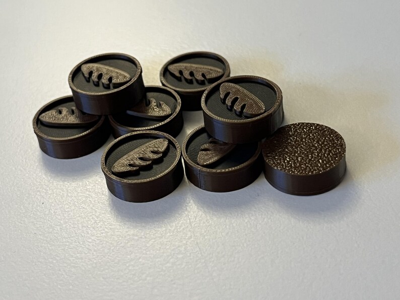 Puede incluir: Una colecci&oacute;n de fichas de juego circulares marrones. Cada pieza tiene un borde marr&oacute;n oscuro, un interior negro y un dise&ntilde;o en relieve marr&oacute;n claro. Una pieza tiene una superficie texturizada.