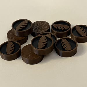 Puede incluir: Una colecci&oacute;n de fichas de juego circulares marrones, con un centro negro y un dise&ntilde;o marr&oacute;n en forma de pan. Algunas piezas tienen una superficie texturizada. Las piezas est&aacute;n esparcidas sobre una superficie blanca.