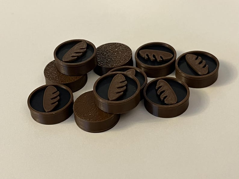 Puede incluir: Una colecci&oacute;n de fichas de juego circulares marrones, con un centro negro y un dise&ntilde;o marr&oacute;n en forma de pan. Algunas piezas tienen una superficie texturizada. Las piezas est&aacute;n esparcidas sobre una superficie blanca.