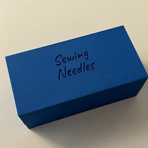 May include: A rectangular, cobalt blue box with the words "Sewing Needles" embossed on the top. The box has a textured surface and is set against a white background. The box is a storage container for sewing supplies.
