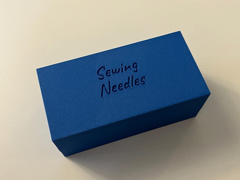 May include: A rectangular, cobalt blue box with the words "Sewing Needles" embossed on the top. The box has a textured surface and is set against a white background. The box is a storage container for sewing supplies.