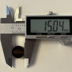Puede incluir: Un calibrador digital mide un peque&ntilde;o bot&oacute;n redondo de color marr&oacute;n oscuro con un dise&ntilde;o de hogaza de pan. El bot&oacute;n mide 15,04 mm de di&aacute;metro. El calibrador tiene una pantalla digital y est&aacute; marcado con "mm/in", "ON/OFF" y "ZERO". El bot&oacute;n mide aproximadamente 0,59 pulgadas.