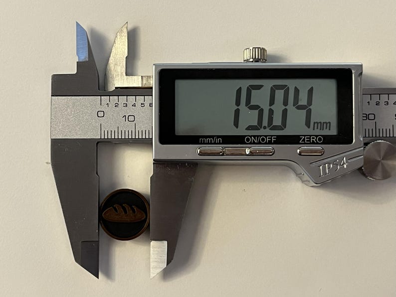 Puede incluir: Un calibrador digital mide un peque&ntilde;o bot&oacute;n redondo de color marr&oacute;n oscuro con un dise&ntilde;o de hogaza de pan. El bot&oacute;n mide 15,04 mm de di&aacute;metro. El calibrador tiene una pantalla digital y est&aacute; marcado con "mm/in", "ON/OFF" y "ZERO". El bot&oacute;n mide aproximadamente 0,59 pulgadas.