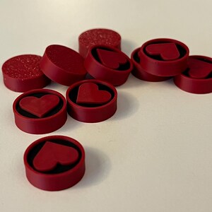 Puede incluir: Un conjunto de tapones de botella rojos con dise&ntilde;os de coraz&oacute;n. Algunos tapones son de color rojo liso, mientras que otros tienen una textura brillante. Los detalles en forma de coraz&oacute;n est&aacute;n en relieve.