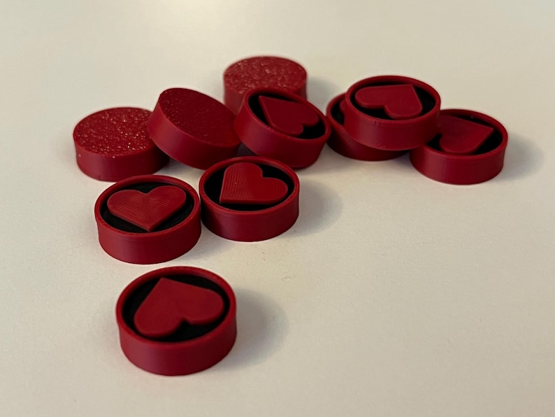 Puede incluir: Un conjunto de tapones de botella rojos con dise&ntilde;os de coraz&oacute;n. Algunos tapones son de color rojo liso, mientras que otros tienen una textura brillante. Los detalles en forma de coraz&oacute;n est&aacute;n en relieve.