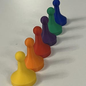 May include: A row of six colorful game pieces in yellow, orange, red, purple, green, and blue. Each piece has a rounded top and a wide base, suitable for board games or educational activities.