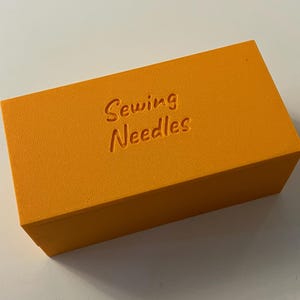 May include: An orange rectangular box with the words "Sewing Needles" embossed on the top. The box is a solid, bright orange color and appears to be made of a textured material. The text is in a cursive font.