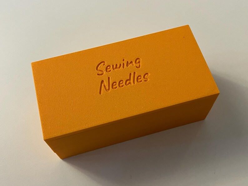 May include: An orange rectangular box with the words "Sewing Needles" embossed on the top. The box is a solid, bright orange color and appears to be made of a textured material. The text is in a cursive font.