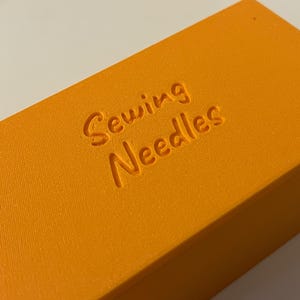 May include: Close-up of an orange rectangular box with the words "Sewing Needles" embossed on the top. The box has a textured surface and is likely used for storing sewing supplies. The color is a vibrant, solid orange.