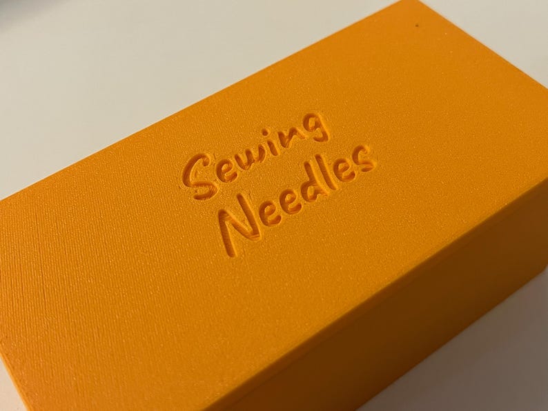 May include: Close-up of an orange rectangular box with the words "Sewing Needles" embossed on the top. The box has a textured surface and is likely used for storing sewing supplies. The color is a vibrant, solid orange.