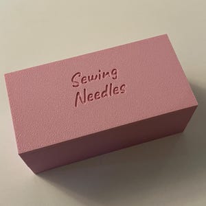 May include: A rectangular, light pink box with the words "Sewing Needles" embossed on the top. The box has a textured surface and is likely used for storing sewing supplies.