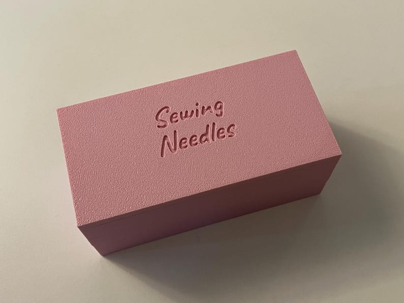 May include: A rectangular, light pink box with the words "Sewing Needles" embossed on the top. The box has a textured surface and is likely used for storing sewing supplies.