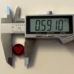 Puede incluir: Un calibrador digital mide un objeto peque&ntilde;o, redondo y negro con un dise&ntilde;o de coraz&oacute;n rojo. El objeto mide aproximadamente 1,5 cm de di&aacute;metro. La pantalla digital del calibrador muestra la medida en pulgadas y mil&iacute;metros.