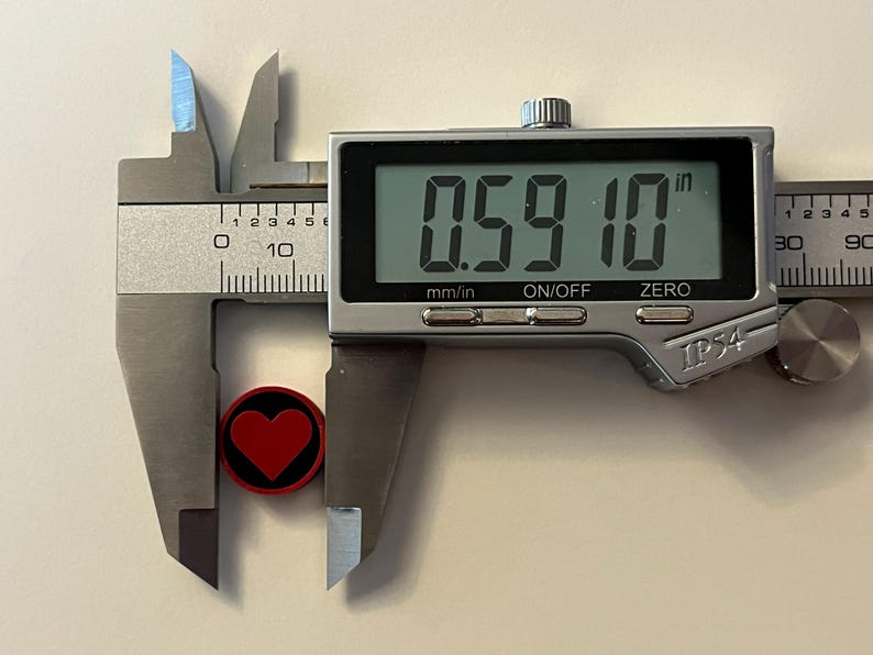 Puede incluir: Un calibrador digital mide un objeto peque&ntilde;o, redondo y negro con un dise&ntilde;o de coraz&oacute;n rojo. El objeto mide aproximadamente 1,5 cm de di&aacute;metro. La pantalla digital del calibrador muestra la medida en pulgadas y mil&iacute;metros.