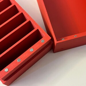 May include: A red wooden organizer with multiple vertical compartments, ideal for storing small items. The organizer is rectangular and has a matching lid. Small silver magnets are visible on the sides of the organizer and lid.