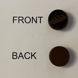 Puede incluir: Dos botones circulares marrones. Un bot&oacute;n tiene un fondo negro con un icono de pan. El otro bot&oacute;n es marr&oacute;n liso. Las palabras "FRONT" y "BACK" est&aacute;n impresas en el fondo blanco.