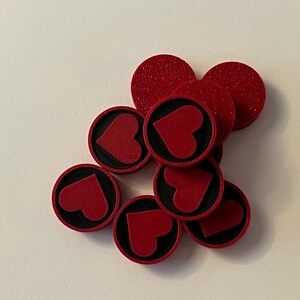 Puede incluir: Un conjunto de fichas de juego rojas y circulares con una superficie texturizada. Cada ficha presenta un borde negro y un dise&ntilde;o de coraz&oacute;n rojo en el centro. Las fichas est&aacute;n dispersas sobre una superficie blanca.