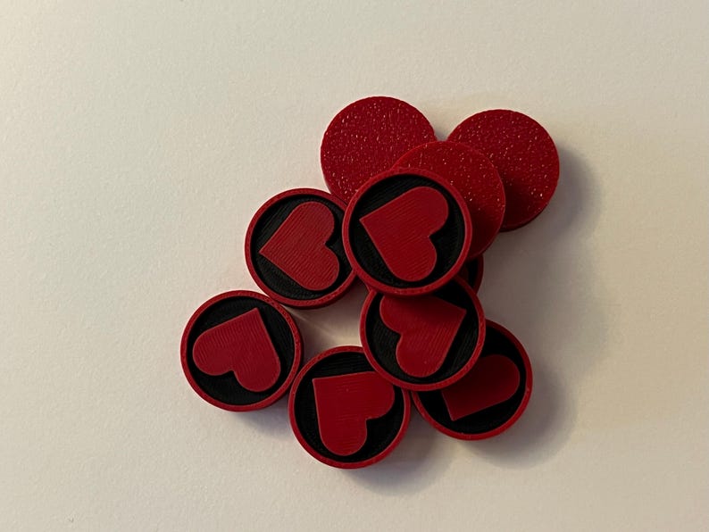 Puede incluir: Un conjunto de fichas de juego rojas y circulares con una superficie texturizada. Cada ficha presenta un borde negro y un dise&ntilde;o de coraz&oacute;n rojo en el centro. Las fichas est&aacute;n dispersas sobre una superficie blanca.