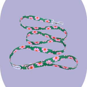 May include: A green dog leash with a repeating pattern of pink daisies. The leash has a silver metal clasp and a matching metal ring. The brand name "pepper grey" is printed on the leash.